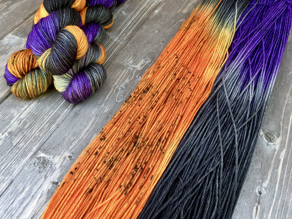 A bold swatch of speckled orange, deep black and rich purple yarn rests beside a stack of three skeins in the upper left corner. The background is a light grey wood.