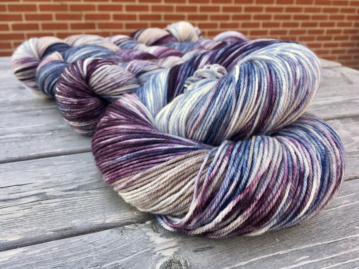 A swirling twist of yarn rests in the foreground, twisted skeins peek out from behind in the background. The yarn is resting against a grey wooden table and a red brick wall is behind everything.