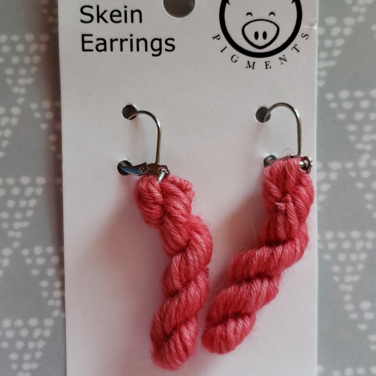 Miniature skeins dangle from surgical steel loops hanging from a white card with the Oink Pigment logo at the top. A dotty triangular pattern on grey can be see in the background.