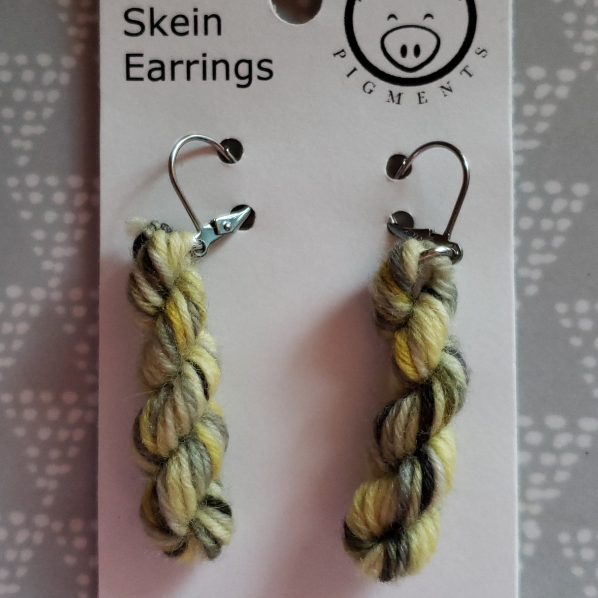 miniature skeins of yarn dangle from steel earring hooks on a white card with the Oink Pigments logo and information at the top.