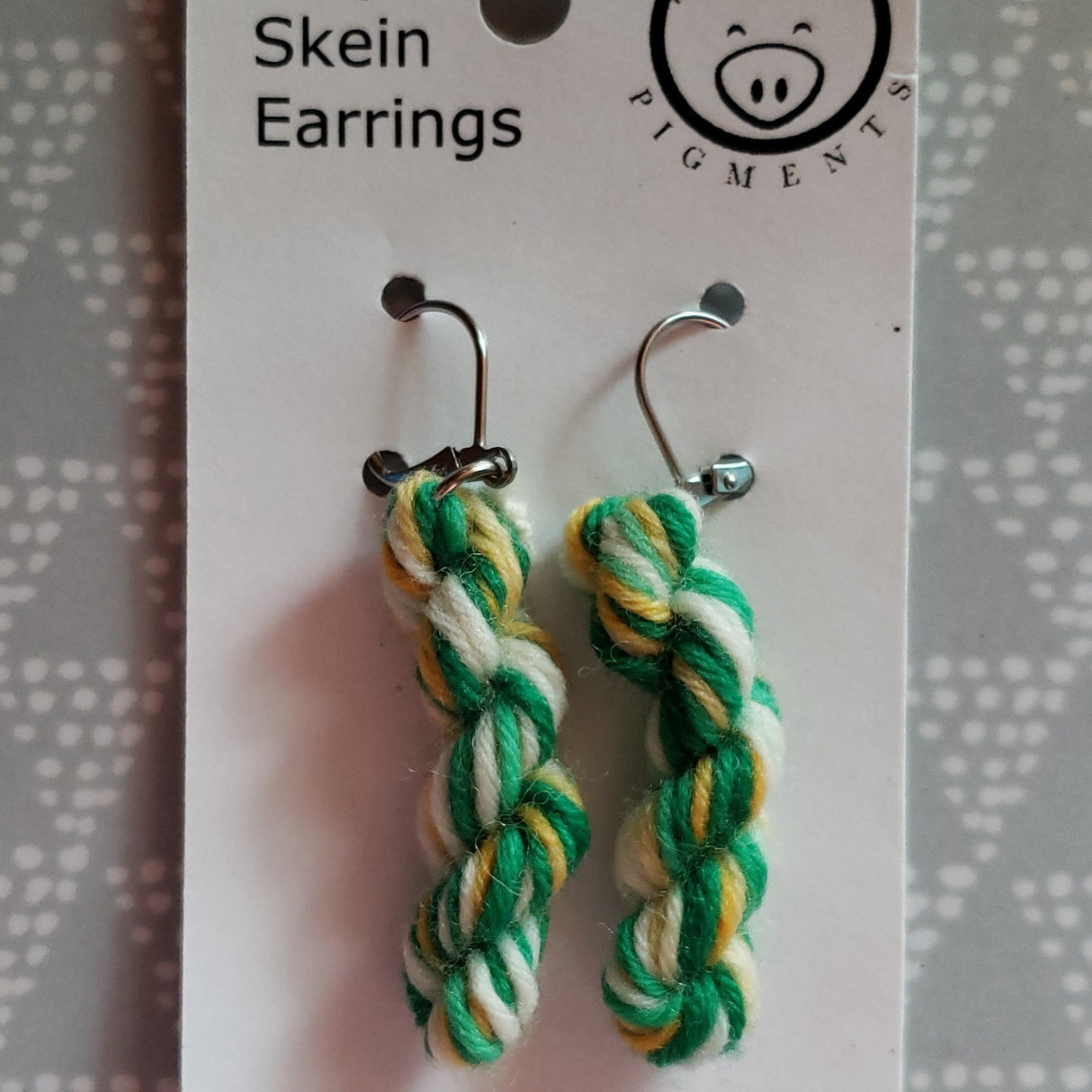 miniature skeins of yarn dangle from steel earring hooks on a white card with the Oink Pigments logo and information at the top.