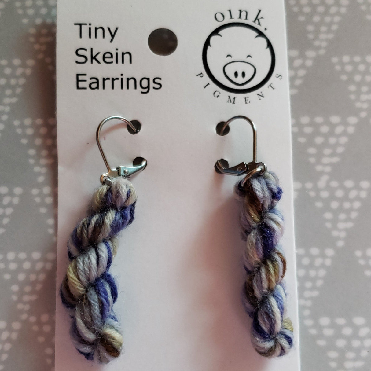 miniature skeins of yarn dangle from steel earring hooks on a white card with the Oink Pigments logo and information at the top.
