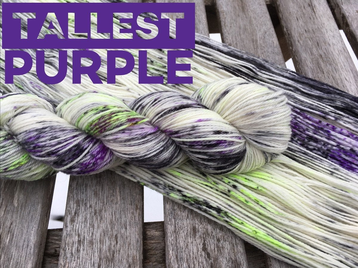a swath of flat yarn sits on a wooden background with a twisted skein of yarn on top. The yarn is snowy white with bright neon lime, deep purple, and black speckles. Purple text in the upper right corner reads "tallest purple"