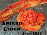 skein of yarn twisted in a spiral and one laid out both are a bright orange with splashes of deep orange and black speckles, black text reads "Spiked cider July 20202"