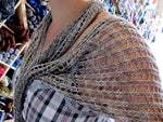 A grey mottled lacy wrap surrounds the shoulders of a light skinned person wearing a plaid dress.  A wall of yarn is in the background. 