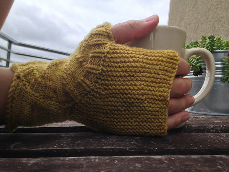 a light skinned hand, clad in a beautiful mustard yellow fingerless mitt, holds a white mug against a dark wooden background. a metal railing and a cloudy sky can be seen in the upper left corner. a beige wall and a small plant rest in the upper right corner. 
