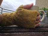 a light skinned hand, clad in a beautiful mustard yellow fingerless mitt, holds a white mug against a dark wooden background. a metal railing and a cloudy sky can be seen in the upper left corner. a beige wall and a small plant rest in the upper right corner. 