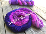 Vivid magenta-pink and deep royal purple swirl in a cinnamon bun shape of yarn that rests on a wooden background.  At the top of the frame is a second twisted skein of yarn.  White text reads "Thistle be perfect" across the middle of the frame. 