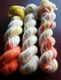 Three skeins showcase the variety of variations that occur from handdying. Color swirls or sparkles differently on each one.  They're resting against a dark maroon background. 