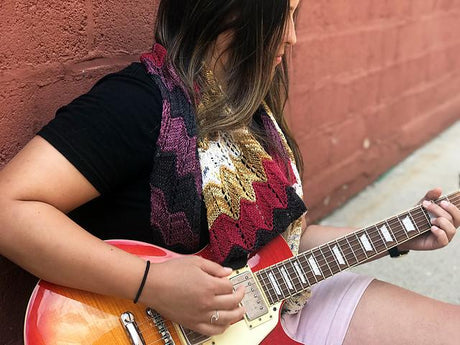 A dark haired person playing guitar against a brick wall, wears a multicolored shawlette.