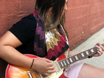 A dark haired person playing guitar against a brick wall, wears a multicolored shawlette.
