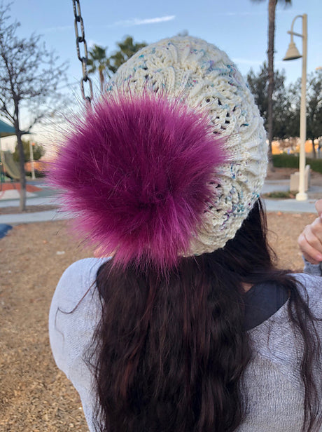 A fluffy fuchsia pompom sits on a knitted lacy hat. There is a playground in the background. 