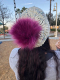 A fluffy fuchsia pompom sits on a knitted lacy hat. There is a playground in the background. 