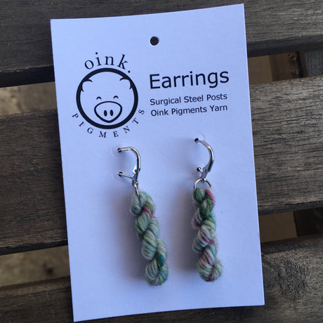 two miniature skeins of yarn on silver colored ear hooks rest on a white card with Oink Pigment logo and information at the top.  The card rests on a slatted dark wooden background.