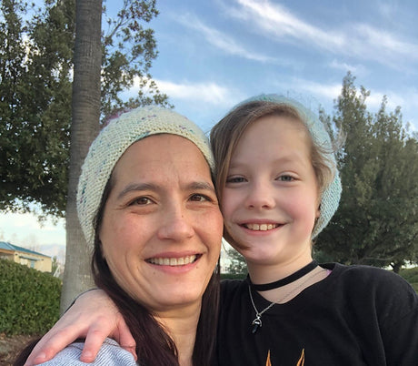 an adult and a child pose for the camera, each wearing knitted hats. They are smiling and have arms around one another. 