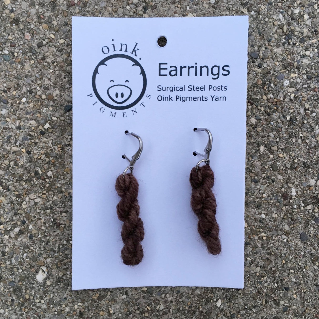 miniature skeins of yarn dangle from steel earring hooks on a white card with the Oink Pigments logo and information at the top.