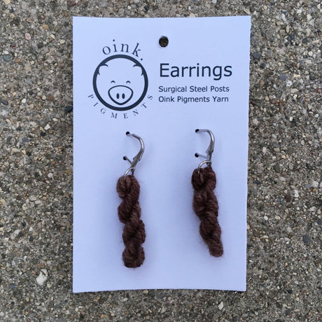 miniature skeins of yarn dangle from steel earring hooks on a white card with the Oink Pigments logo and information at the top.