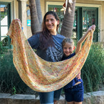 A smiling adult stands next to a smiling child while they each hold one end of a beautiful lacy golden speckled shawl.