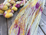 A river of colorful yarn swoops diagonally across the frame, a stack of three skeins rest in the upper left of the frame.  The yarn is resting on a grey wooden background. 