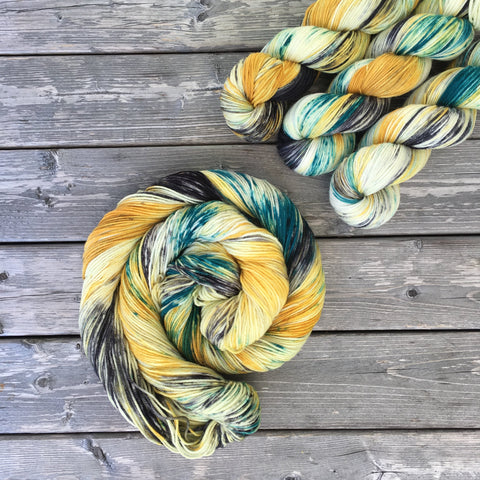 A colorful swirl of yarn curls around itself while three twisted skeins rest in the upper right corner.  The yarn is various deep yellow and green tones. 
