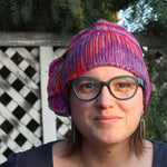 A light skinned femme person wearing green glasses gazes at the camera with a serene expression on their face. they are sporting a brightly colored ruffled knitted hat.