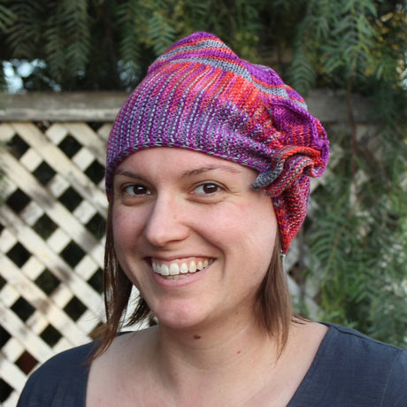a light skinned femme person sports a ruffled and brightly colored hat.  They are smiling at the camera.  There is a white fence and green foliage in the background.