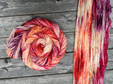 A swirl of yarn rests to the left of a unhanked skein.  Both skeins rest on a wooden background.