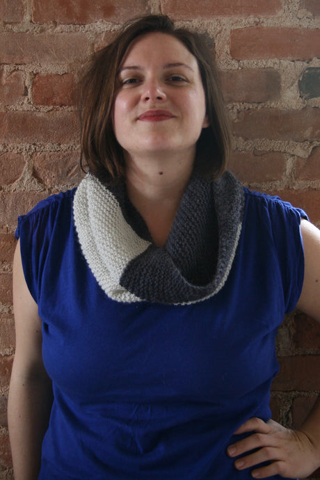 A confident, smiling person in a royal blue top wears a bold, graphic cowl in dark charcoal and cream.  They are standing in front of a brick background. 