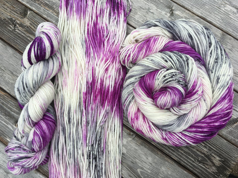 Three skeins, in three different arrangements (L to R: a doubled over twist, spread flat, and swirled around itself) rest against a grey wooden background.