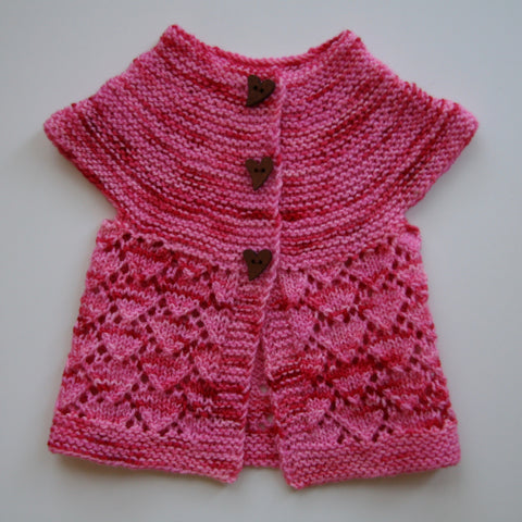 A petite pink and sweater with red streaks sits atop a white background. The top of the little sweater is in a swooping garter stitch, and the body has a delicate lacy heart pattern.  Three dark heart buttons complete the darling look. 