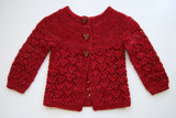 A textured red sweater rests on a clean white background.  The yoke of the sweater is garter stitch and the sleeves and body are a delicate heart lace pattern.  Three heart shaped buttons complete the sweetness. 