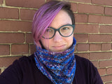 A softly smiling light skinned person stands against a brick wall modeling a textured cowl.  Their pinky-purple hair and dark glasses set the yarn off perfectly. 
