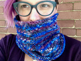 A lovely rose-haired person peeks out fromn behind a richly textured cowl.  They are wearing dark glasses and a deep purple top. 