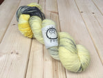 a pale cool yellow skein with a smudge of charcoal and deeper yellow rests on a pale wooden background.