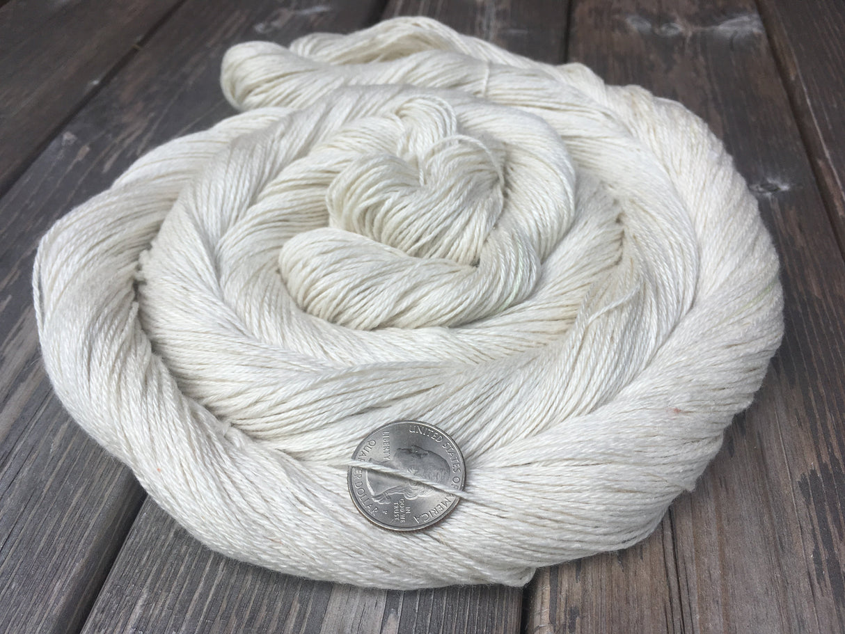 A shimmery swirl of creamy un-dyed yarn rests on a dark wooden background. A US quarter rests behind on strand to showcase the thickness.