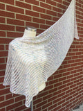 A pale toned lacy wrap is artfully draped over a white mannequin and up onto a brick background. 