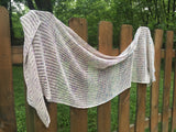 A lacey wrap with an open pattern and strong lines is draped over a wooden fence.  There is greenery in the background. 