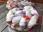 A deliciously fluffy braid of pale fiber with splashes of plummy purple, lobster red, and grey rests on a cool grey wooden surface. 