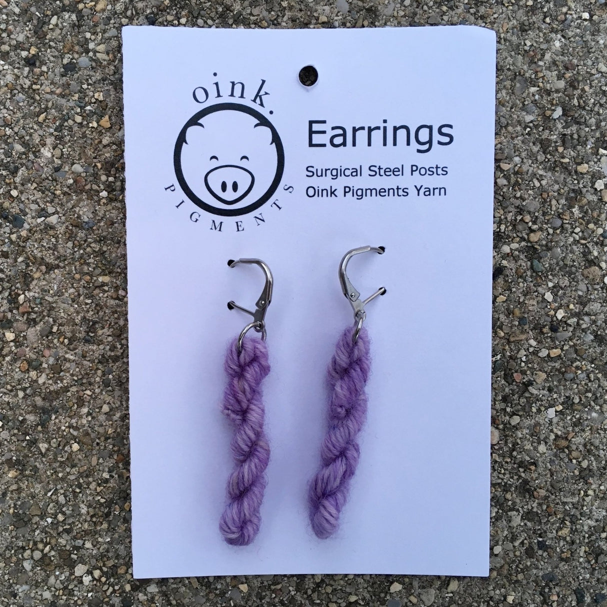 miniature skeins of yarn dangle from steel earring hooks on a white card with the Oink Pigments logo and information at the top.