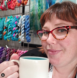 A beautiful person with fabulous glasses holds a white mug with a teal interior. Behind them is a wall of brightly colored yarn. 