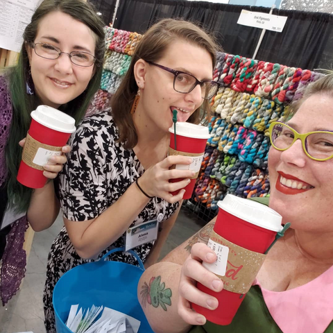 Three smiling faces of Team Oink against a wall of dazzling yarn.  Each person is holding a red coffee cup.