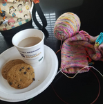 A picture perfect pause; two cookies and a cup of coffee rest on a white paper plate surrounded by a colorful project bag and a sock in progress. 