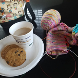 A picture perfect pause; two cookies and a cup of coffee rest on a white paper plate surrounded by a colorful project bag and a sock in progress. 
