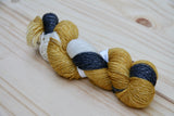 A golden, cream, and black skein of yarn with a subtle glimmer rests on a pale wooden background. 