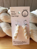 miniature skeins of yarn dangle from steel earring hooks on a white card with the Oink Pigments logo and information at the top. The card is resting against white skeins of yarn.