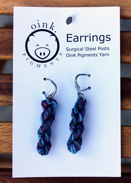 miniature skeins of yarn dangle from steel earring hooks on a white card with the Oink Pigments logo and information at the top.