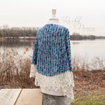 A beautiful shawl with strong vertical ridges and a delicate cream crocheted lace rests on a mannequin in a restful lakeside location.