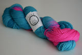 A twisted skein of intense pink and teal  rests against a pale wooden background. 