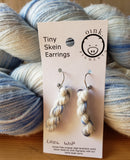 miniature skein earrings rest on a white card with the Oink Pigments logo and text at the top of the card. The card is leaning against two large skeins of blue and white yarn.
