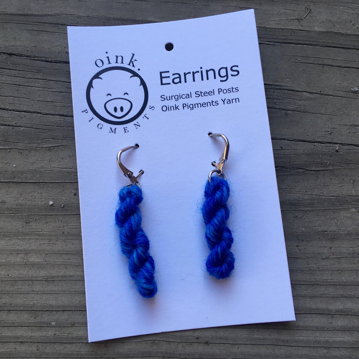 miniature skeins of yarn dangle from steel earring hooks on a white card with the Oink Pigments logo and information at the top.